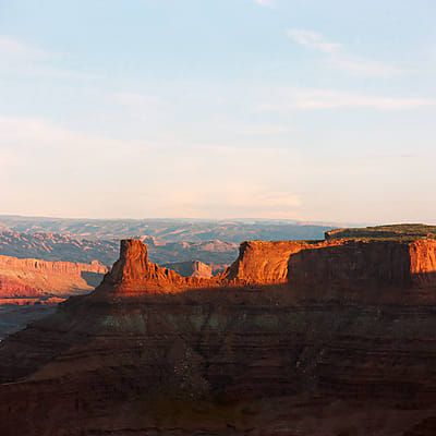 Mesa formation rising above a canyon at first light, illuminated against a wide pale sky.